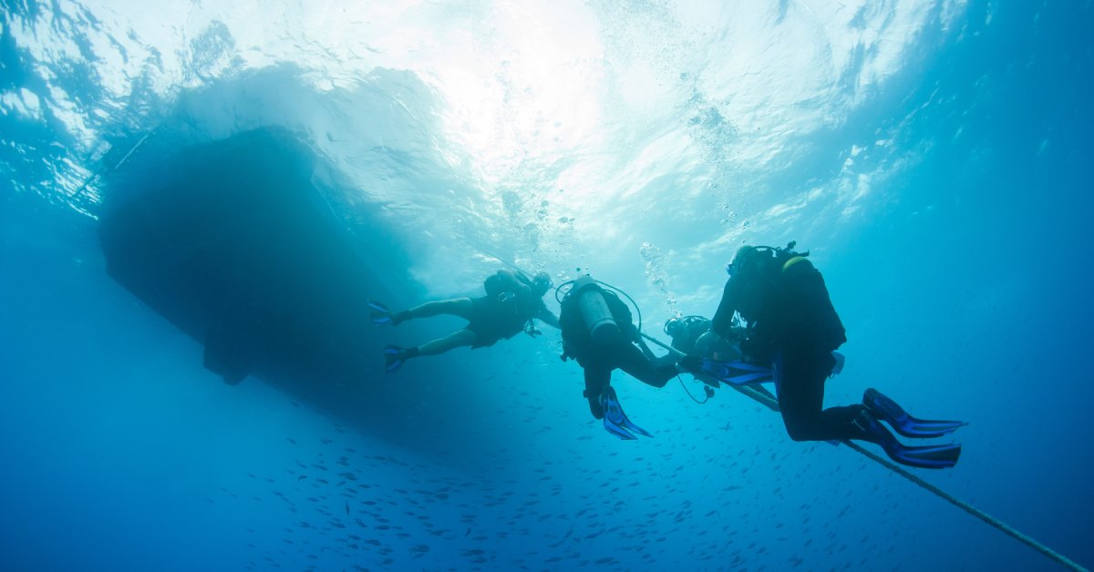 Four scuba divers underwater, decompressing as they hold onto a rope connected to a ship overhead.