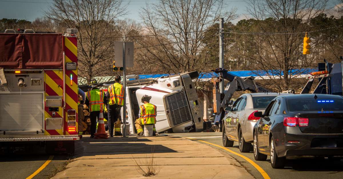 A massive logging truck is overturned in the middle of the road, and emergency crews are gathering around to help.