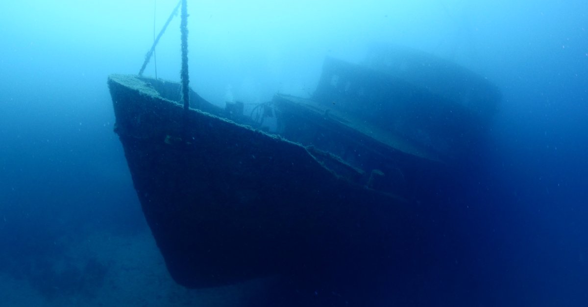 A large sunken boat deep underwater. The water is murky, and sunlight is barely visible above.